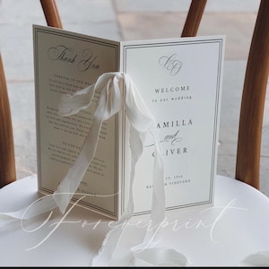 K&ouml;nnte beinhalten: Ein cremefarbenes Hochzeitsprogramm mit schwarzem Text, gebunden mit einem wei&szlig;en Band. Der Text enth&auml;lt "Danke" und "Willkommen zu unserer Hochzeit". Die Namen "Amilla und Oliver" sind ebenfalls sichtbar.