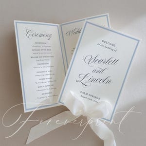 May include: Wedding ceremony programs with a light blue border and white ribbon. The programs are printed with elegant calligraphy, including the names "Scarlett and Lincoln" and the date "August 25th, 20xx". The program lists the ceremony details.