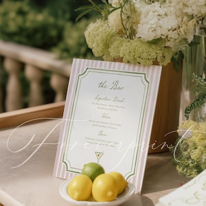May include: A bar menu sign with pink and white stripes and green trim. The sign lists signature drinks, wine, and beer options. A small white dish holds lemons and a lime. Floral arrangements are in the background.