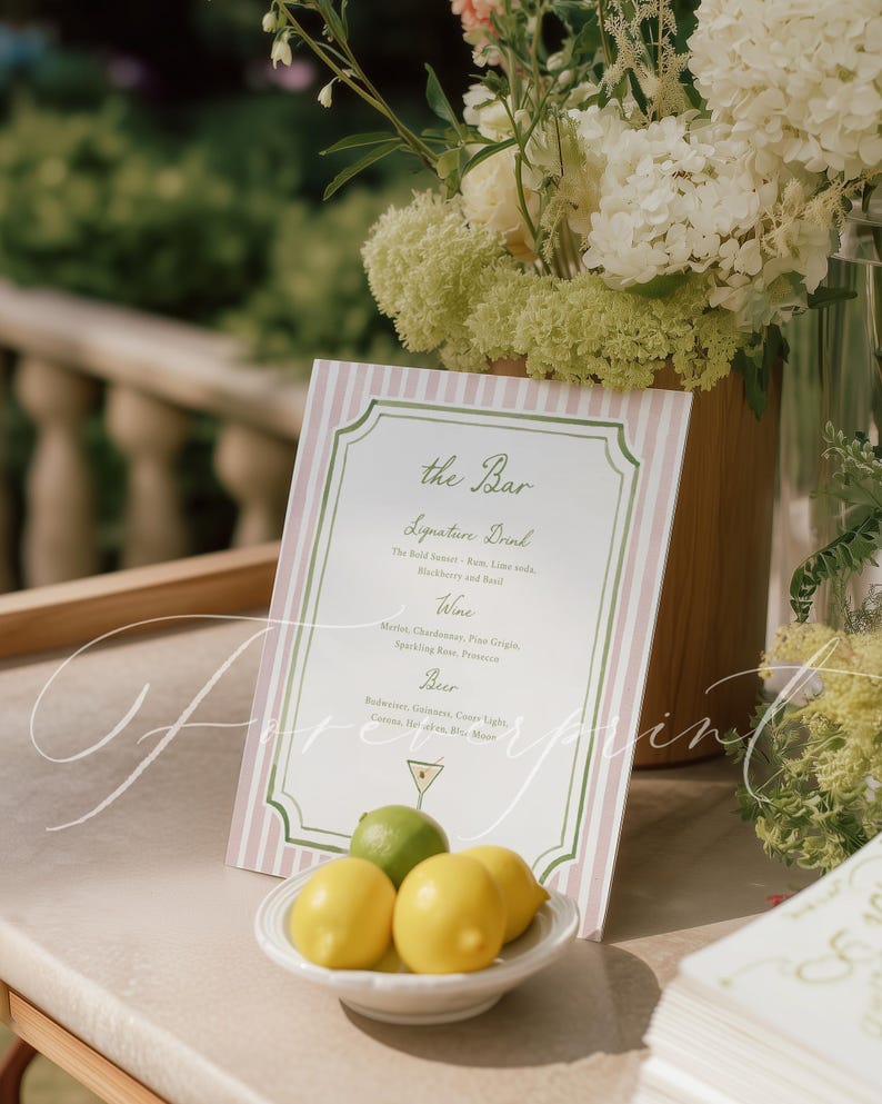 May include: A bar menu sign with pink and white stripes and green trim. The sign lists signature drinks, wine, and beer options. A small white dish holds lemons and a lime. Floral arrangements are in the background.