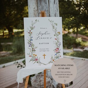 May include: A white sign with a floral wreath border and the text "Welcome to Sophie Linnea's Baptism Cleveland, Ohio 14th August 2029" with a gold cross. The sign is on an easel with a brown wooden base.