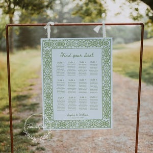 May include: A wedding seating chart with the text "Find your Seat" in elegant script. The chart features a green floral border and lists names under table numbers. The sign is suspended from a copper-colored metal frame.