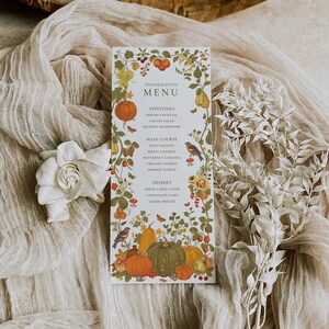 May include: A Thanksgiving menu card with a white background and a floral border. The menu includes appetizers, main course, and dessert options. The card is decorated with pumpkins, gourds, and other fall foliage.