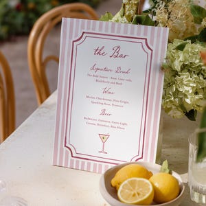Peut inclure: Un menu de bar à rayures roses et blanches avec une bordure décorative. Le menu répertorie les cocktails signatures, les vins et les bières. Un petit bol de citrons est placé devant le menu, avec une illustration de verre à cocktail.
