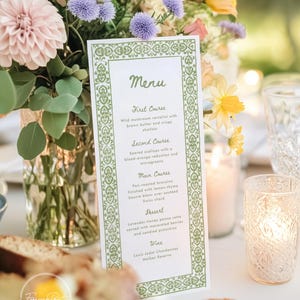 Puede incluir: Una tarjeta de menú con un borde decorativo verde, que detalla una comida de varios platos. El menú incluye platos como raviolis de champiñones silvestres y bronzino asado. La tarjeta está sobre una mesa con flores y velas.