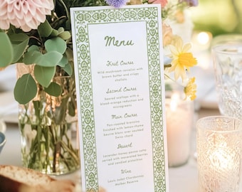 May include: A printed menu card with a green decorative border, listing a multi-course meal. The menu includes dishes like wild mushroom ravioli and pan-roasted bronzino. The card is set on a table with flowers and candles.