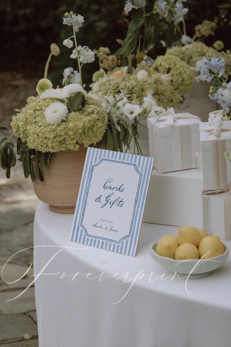 Pu&ograve; includere: Un'impostazione della tavola con un cartello che recita "Cards & Gifts" in blu su sfondo a righe blu e bianche. Una ciotola di limoni &egrave; posta vicino al cartello. Sono presenti anche scatole regalo bianche e composizioni floreali.