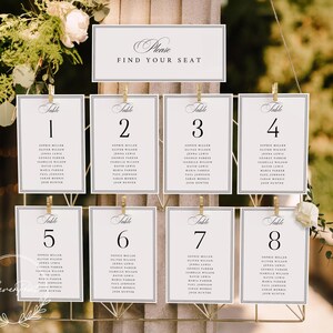 May include: Elegant wedding seating chart with eight table cards, each numbered and listing guest names. A top sign reads "Please Find Your Seat." The cards are held by gold clips on a decorative metal frame, adorned with floral accents.