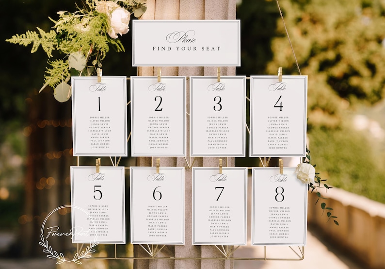 May include: Elegant wedding seating chart with eight table cards, each numbered and listing guest names. A top sign reads "Please Find Your Seat." The cards are held by gold clips on a decorative metal frame, adorned with floral accents.