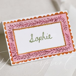 May include: A folded place card with a pink and orange scalloped border. The inner frame is decorated with a leaf design. The name "Sophie" is written in green script in the centre of the card.