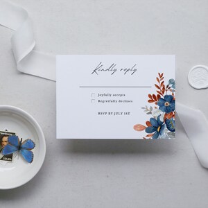May include: A white RSVP card with floral accents in blue, rust, and gray, featuring the text "Kindly reply." A white ribbon and a small bowl with a blue butterfly and a stamp are also present.