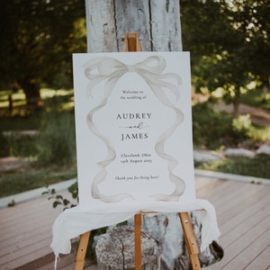 May include: A white wedding sign with a watercolor illustration of a large bow. The sign reads "Welcome to the wedding of Audrey and James, Cleveland, Ohio, 14th August 2023. Thank you for being here!"