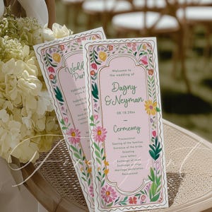 May include: Two pink wedding ceremony programs with floral borders and the text "Welcome to the wedding of Dagny & Neyman." The programs are on a wicker chair with a bouquet of white flowers.