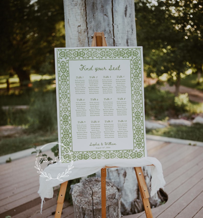 May include: A white framed seating chart with green decorative border and text "Find your Seat". The chart is displayed on a wooden easel, with a white fabric draped below. The names are listed by table number.