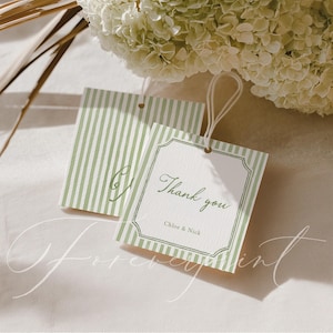 Op de afbeelding: Twee cadeaulabels met een groen en wit gestreept ontwerp. Eén label heeft de woorden "Thank you" en de namen "Chloe & Nick" erop gedrukt. De labels zijn vastgebonden met wit touw en liggen naast een boeket witte bloemen.