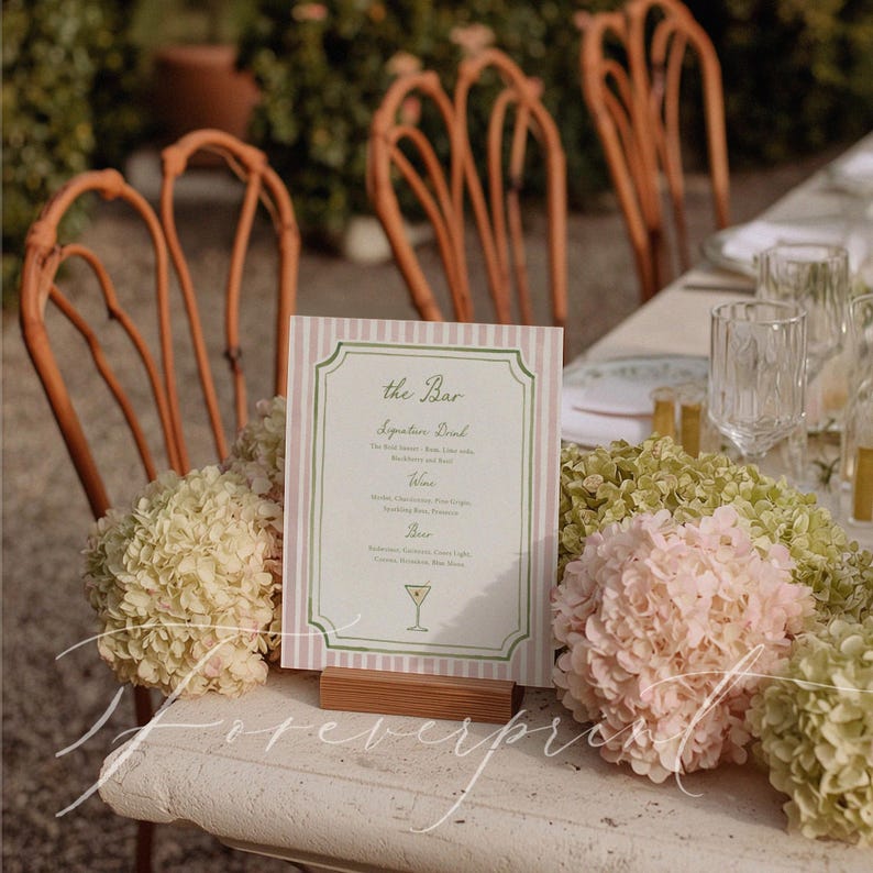 May include: A bar menu with a pink and white striped background and green border sits on a wooden stand. The menu lists signature drinks, wine, and beer. Hydrangea flowers in shades of pink and green surround the menu.