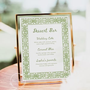 May include: A gold-framed sign with a green border and the text "Dessert Bar" in cursive. The sign lists three dessert options: Wedding Cake, Caramel Bliss, and Sophie's favorite.