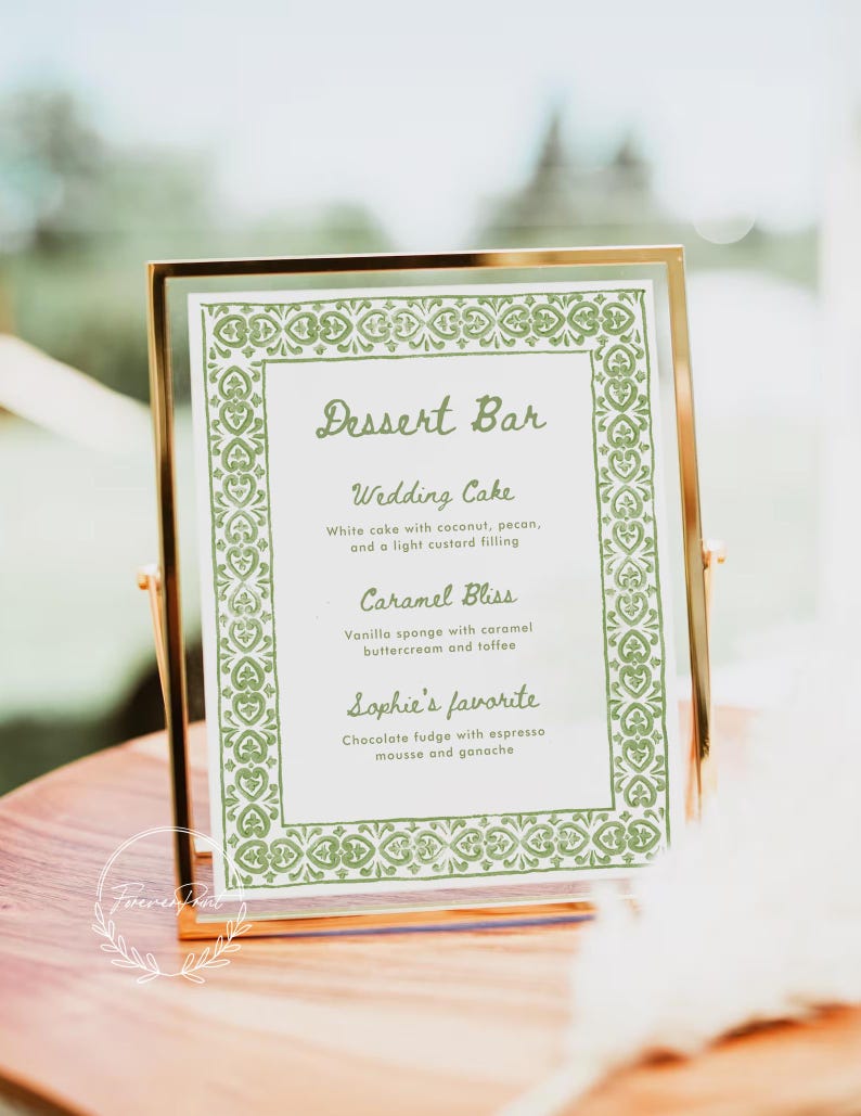 May include: A gold-framed sign with a green border and the text "Dessert Bar" in cursive. The sign lists three dessert options: Wedding Cake, Caramel Bliss, and Sophie's favorite.