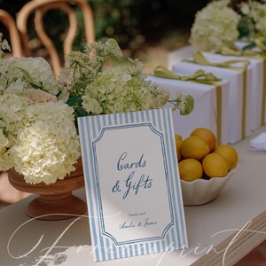 Pu&ograve; includere: Un tavolo da ricevimento nuziale con un cartello con la scritta "Cards & Gifts" in blu, incorniciato da strisce blu e bianche. Sono presenti anche regali bianchi con nastri verdi, una ciotola di limoni e fiori bianchi.