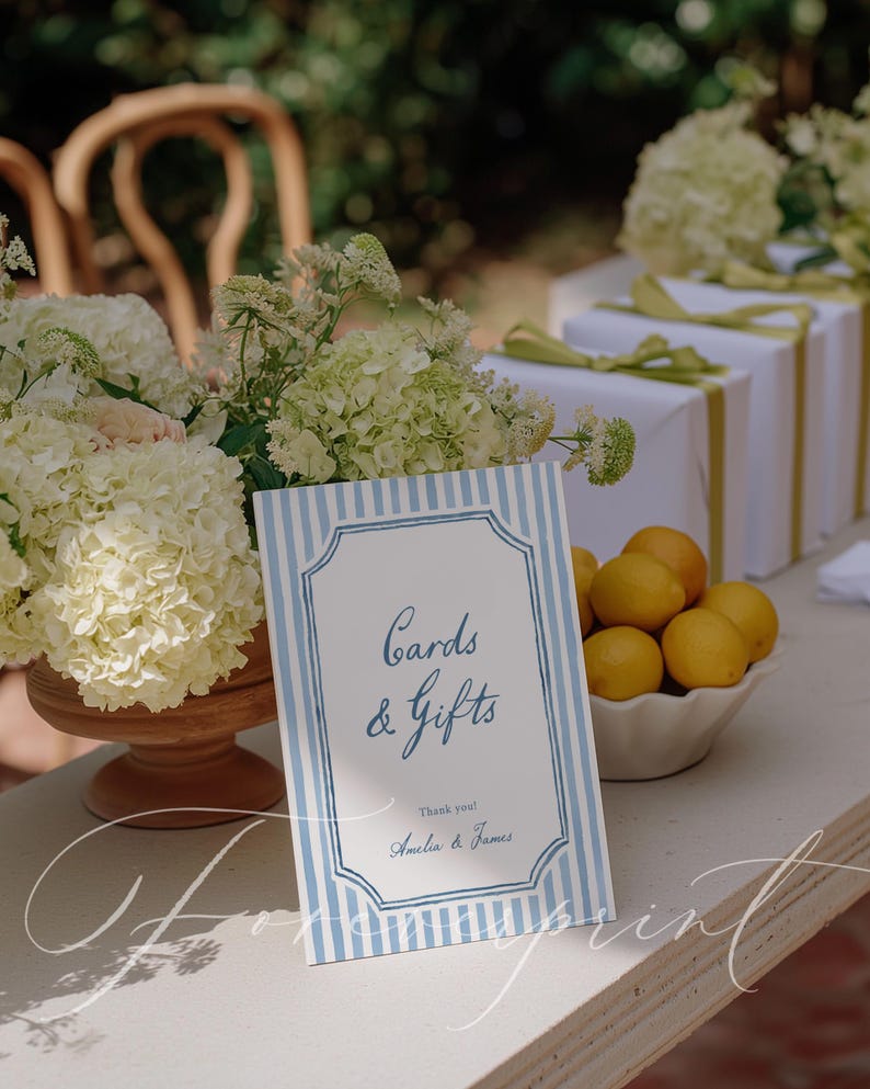 Pu&ograve; includere: Un tavolo da ricevimento nuziale con un cartello con la scritta "Cards & Gifts" in blu, incorniciato da strisce blu e bianche. Sono presenti anche regali bianchi con nastri verdi, una ciotola di limoni e fiori bianchi.