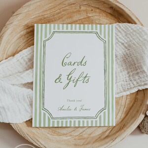 May include: A rectangular sign with green vertical stripes and a decorative border, reading "Cards & Gifts" in elegant script. The sign includes the names "Amelia & James" and is set on a wooden tray with a white cloth.