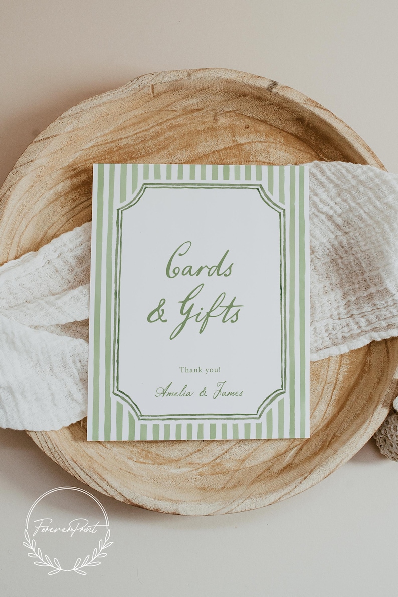 May include: A rectangular sign with green vertical stripes and a decorative border, reading "Cards & Gifts" in elegant script. The sign includes the names "Amelia & James" and is set on a wooden tray with a white cloth.
