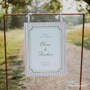 Może przedstawiać: Witamy znak weselny z różowo-białym paskiem w tle. Znak ma dekoracyjną zieloną obwódkę i zawiera imiona "Olivia & Theodore" z lokalizacją i datą.