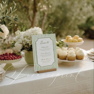 May include: A table setting with a white tablecloth, featuring a dessert menu with green stripes, cupcakes, raspberries, and lemons. The menu lists dessert options. White flowers and greenery add to the elegant display.