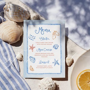 May include: A menu card with a blue border and handwritten text, featuring a menu for a meal. The menu includes a starter, main course, and dessert, with illustrations of seashells, starfish, and lemons. The card is surrounded by seashells on a sandy surface.