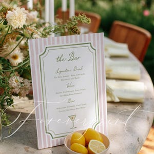 May include: A pink and white striped bar menu with a green border and elegant script. The menu lists signature drinks, wine, and beer options. A small bowl of lemons sits in front of the menu, with floral arrangements in the background.