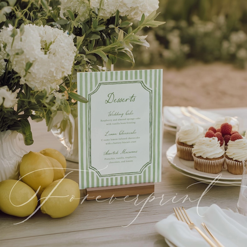 May include: A table setting with a green and white striped menu card that reads "Desserts". The menu is in front of a bouquet of white flowers, lemons, and cupcakes with raspberries. The menu is held in a wooden stand.