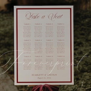 Puede incluir: Un cartel de distribución de mesas de boda blanco con borde rojo oscuro, que presenta el texto Take a Seat, doce asignaciones de mesa y los nombres Scarlett & Lincoln con la fecha August 21, 20xx. Una cinta roja oscura rodea el caballete de madera.