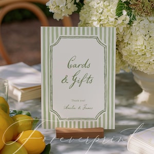 以下が含まれることがあります： 「Cards & Gifts」と書かれたサイン。緑と白のストライプのボーダーで囲まれています。木製の台座に立てられており、「Amelia & James」と「Thank you!」の名前がメインテキストの下にあります。背景にはレモンと花があります。