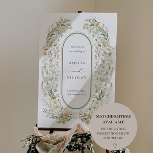 以下が含まれることがあります： 花輪のデザインが施された結婚式のウェルカムサイン。サインには「AmeliaとNicolayの結婚式へようこそ、クリーブランド、オハイオ州、2023年8月14日」と書いてあります。