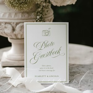May include: A white Photo Guestbook sign with a green border and text. The sign features a camera icon and elegant script that reads "Photo Guestbook". The text below says "Take a photo, place it in the book and leave a message for SCARLETT & LINCOLN."