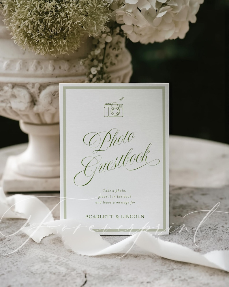 May include: A white Photo Guestbook sign with a green border and text. The sign features a camera icon and elegant script that reads "Photo Guestbook". The text below says "Take a photo, place it in the book and leave a message for SCARLETT & LINCOLN."