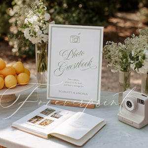 May include: A wedding photo guestbook setup with a sign that reads "Photo Guestbook". The sign is next to an open guestbook with photos and a white instant camera. The table also has lemons and flowers.