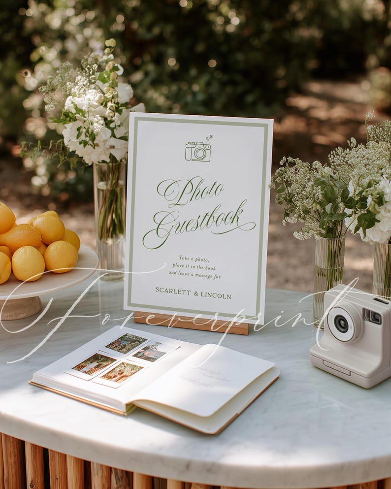 May include: A wedding photo guestbook setup with a sign that reads "Photo Guestbook". The sign is next to an open guestbook with photos and a white instant camera. The table also has lemons and flowers.