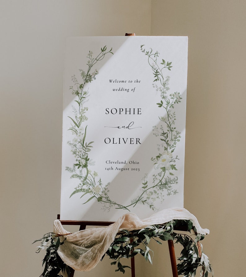 Pode incluir: Um cartaz de casamento branco com um design de coroa de flores. O cartaz diz "Bem-vindos ao casamento de Sophie e Oliver, Cleveland, Ohio, 14 de agosto de 2023".