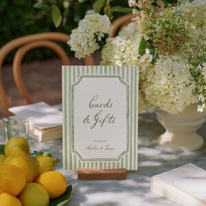 May include: A sign with green and white stripes that reads "Cards & Gifts" with the names "Amelia & James" below. The sign is on a wooden stand, with lemons and flowers in the background.