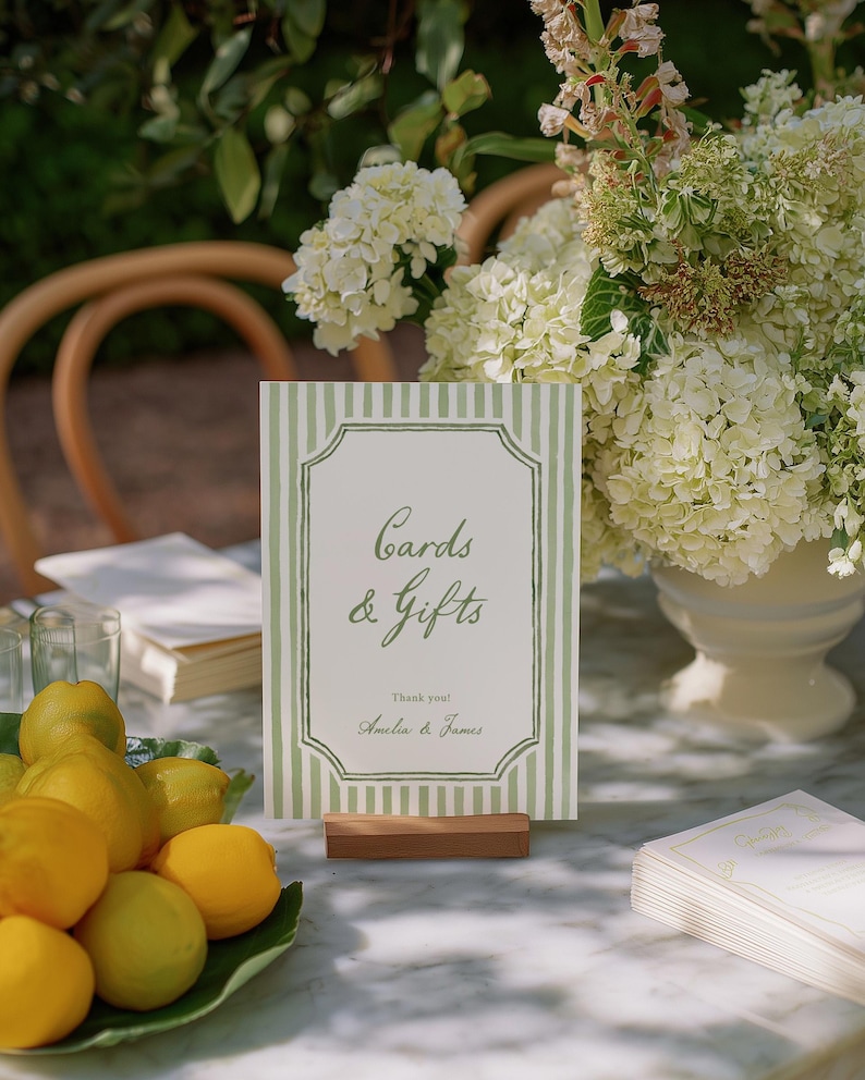 May include: A sign with green and white stripes that reads "Cards & Gifts" with the names "Amelia & James" below. The sign is on a wooden stand, with lemons and flowers in the background.