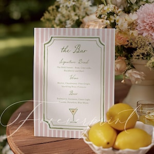 May include: A pink and white striped bar menu with green trim, listing signature drinks, wine, and beer options. A bowl of yellow lemons sits on a wooden table, with a floral arrangement in the background.