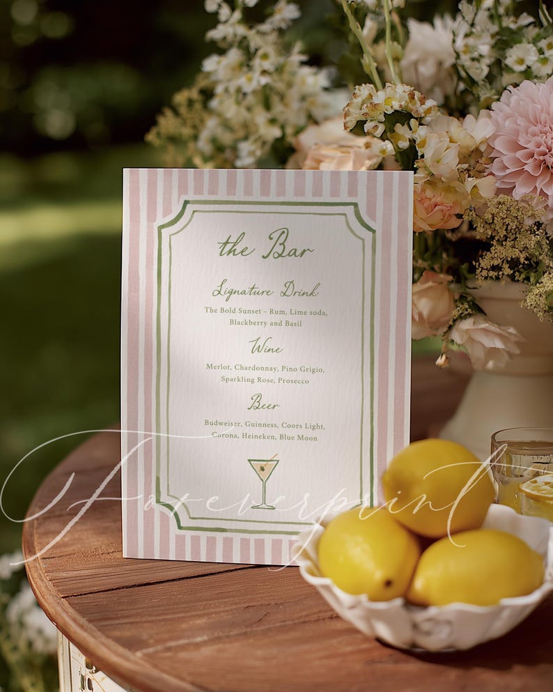 May include: A pink and white striped bar menu with green trim, listing signature drinks, wine, and beer options. A bowl of yellow lemons sits on a wooden table, with a floral arrangement in the background.