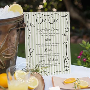 Könnte beinhalten: Ein grün-weiß gestreiftes Schild mit der Aufschrift "Cin Cin". Das Schild listet ein Signature-Getränk, Weine, Biere und alkoholfreie Getränke auf. Ein Eimer mit Eis und ein Glas Getränk sind im Vordergrund.