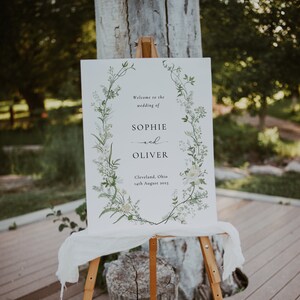 Pode incluir: Um cartaz de casamento branco com uma borda de coroa de flores. O cartaz diz "Bem-vindos ao casamento de Sophie e Oliver, Cleveland, Ohio, 14 de agosto de 2023".