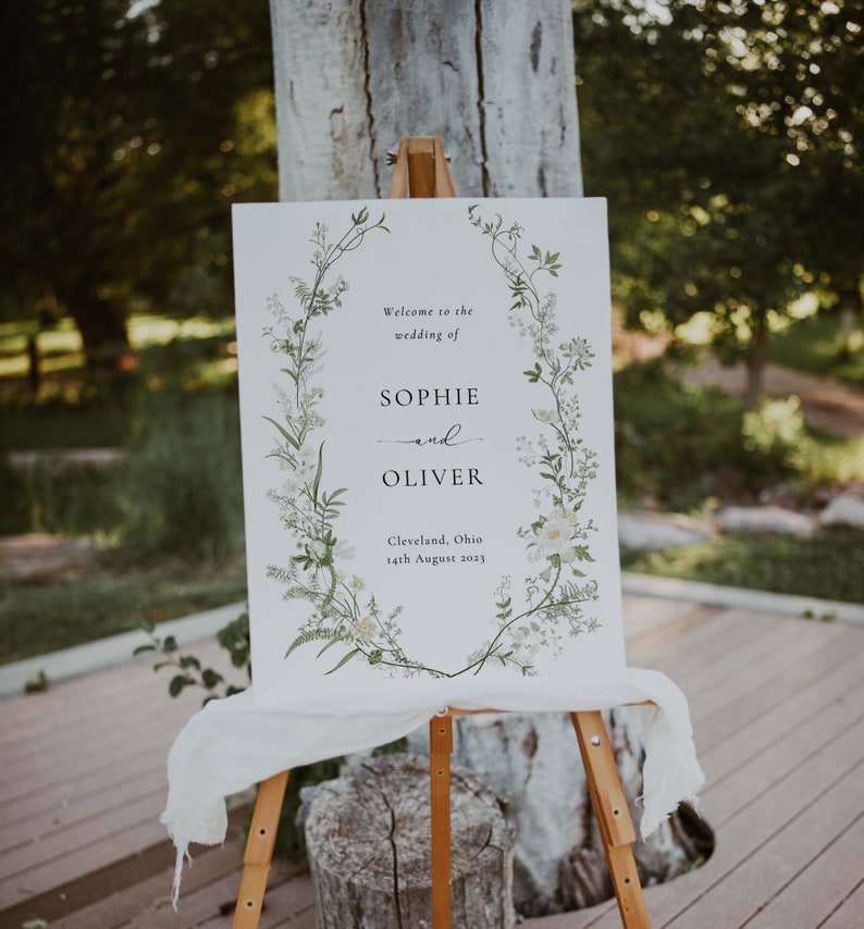 Pode incluir: Um cartaz de casamento branco com uma borda de coroa de flores. O cartaz diz "Bem-vindos ao casamento de Sophie e Oliver, Cleveland, Ohio, 14 de agosto de 2023".