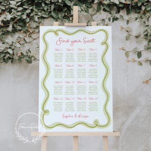 May include: A wedding seating plan displayed on a wooden easel. The white sign features a green and pink design with the words "Find your Seat". Guest names are listed under table numbers, with a decorative border.