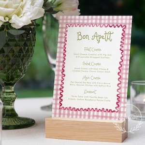 Op de afbeelding: Een menukaart met een roze ruitpatroon en een decoratieve rand, gehouden in een houten standaard. Het menu bevat de woorden "Bon Appetit" en een lijst met gangen. Een groen glas en witte bloemen staan op de achtergrond.