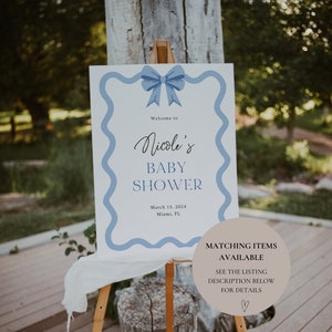 Peut inclure: Une pancarte blanche avec une bordure ondul&eacute;e bleue et un n&oelig;ud bleu sur le dessus. La pancarte indique "Bienvenue &agrave; la Baby Shower de Nicole" avec la date "15 mars 2024" et le lieu "Miami, FL". Un cercle brun avec du texte blanc indique "Articles assortis disponibles" et "Voir la description de la liste ci-dessous pour plus de d&eacute;tails".