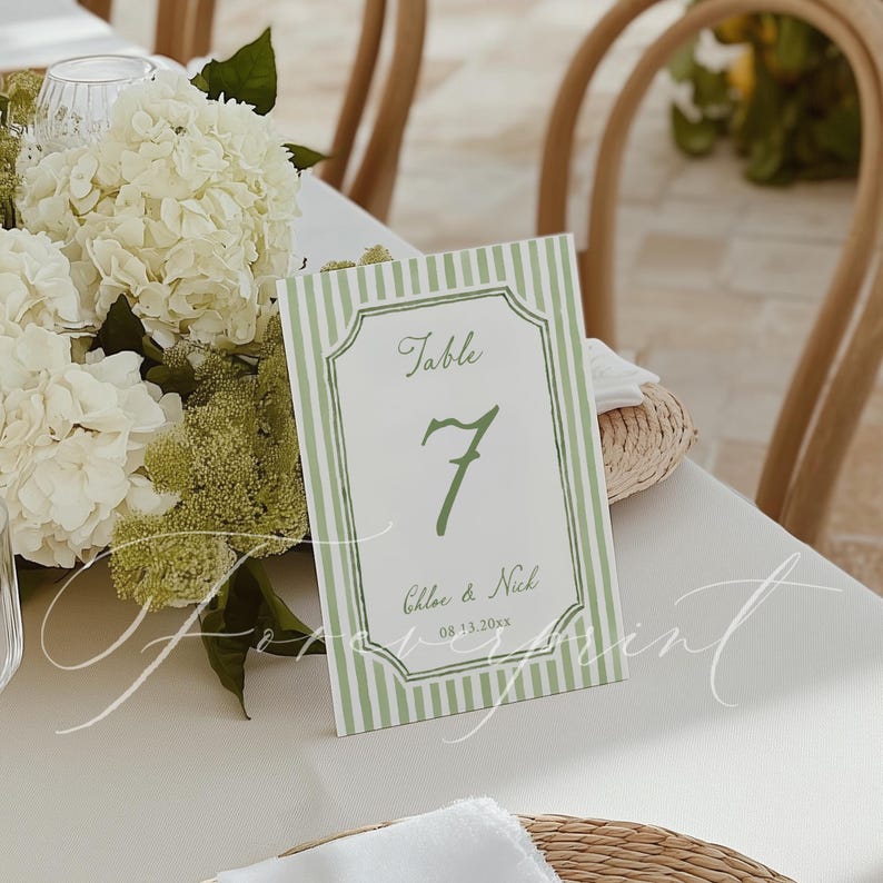 May include: A table number card with a green and white striped border. The card features the word "Table" at the top, the number "7" in the center, and the names "Chloe & Nick" with a date below. The card is on a white tablecloth.